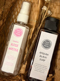 Two skincare products from Forever Earth Skincare arranged in a woven brown basket: a clear spray bottle of Organic Rose Water with a pink label and a dark bottle of African Black Soap with a black cap and white label, accented with dried white flowers.