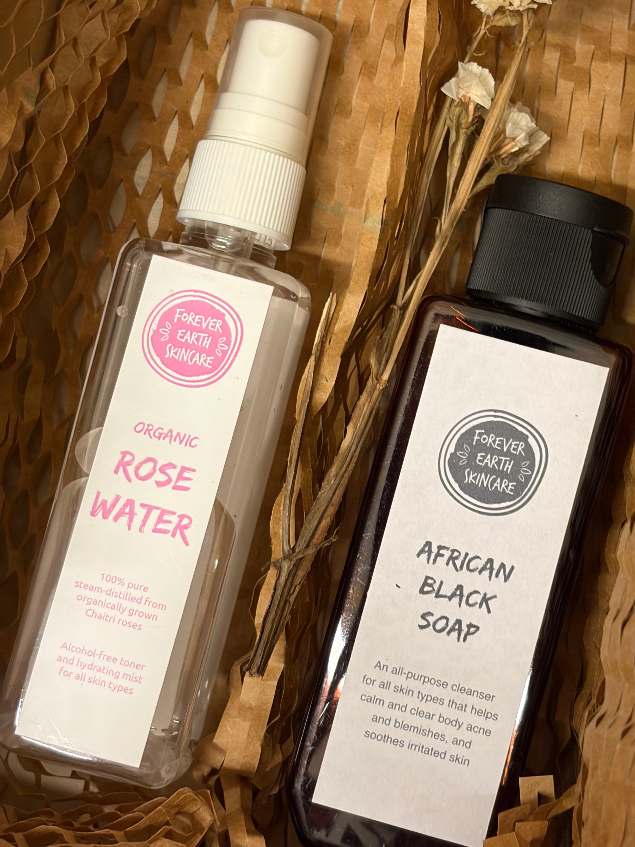 Two skincare products from Forever Earth Skincare arranged in a woven brown basket: a clear spray bottle of Organic Rose Water with a pink label and a dark bottle of African Black Soap with a black cap and white label, accented with dried white flowers.