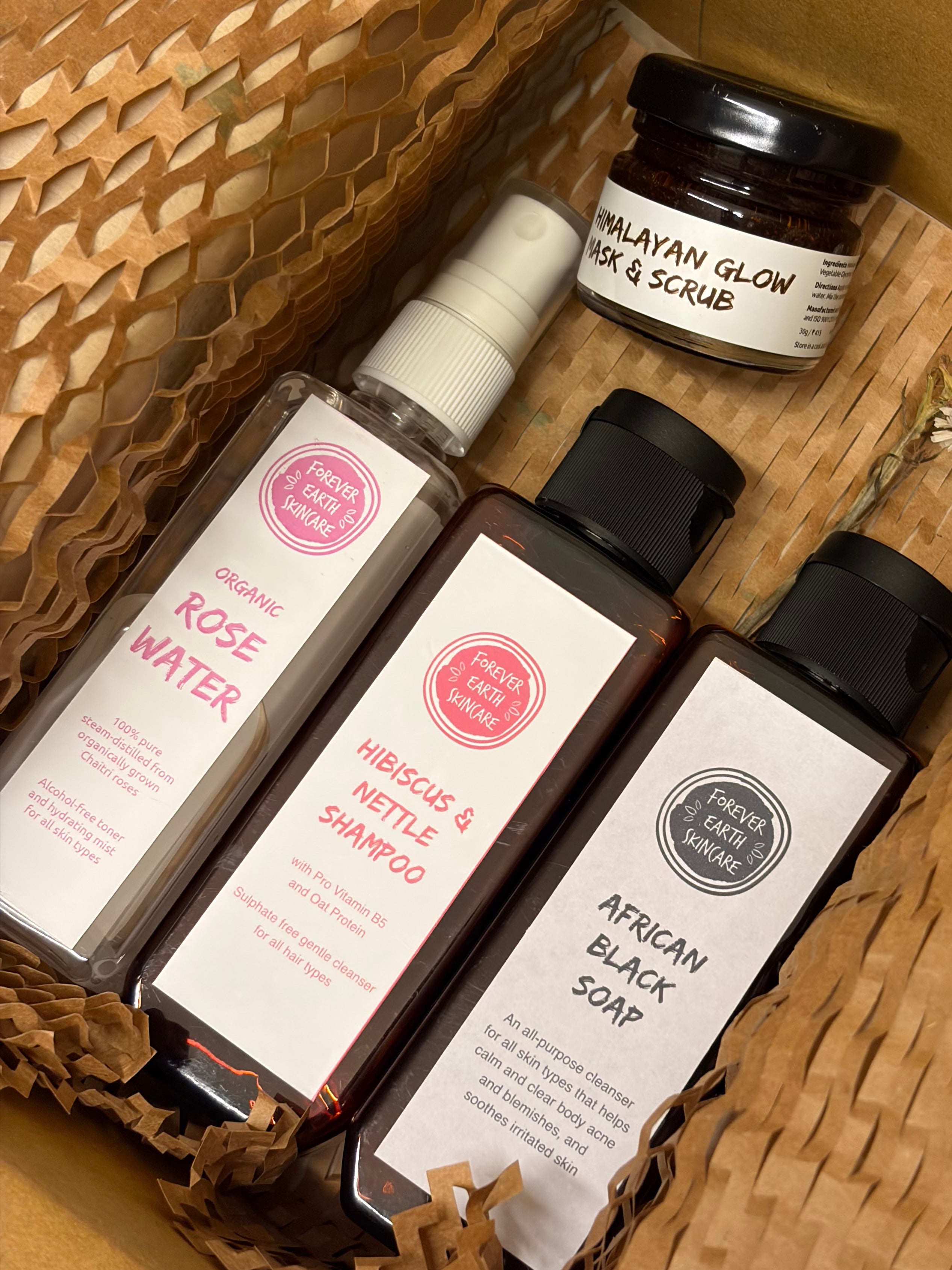 A curated haircare gift set from Hair Care Heroes, including Organic Rose Water, Hibiscus & Nettle Shampoo, African Black Soap, and Malayan Glow Hair & Scrub, arranged in a brown paper honeycomb box with a dried flower sprig.