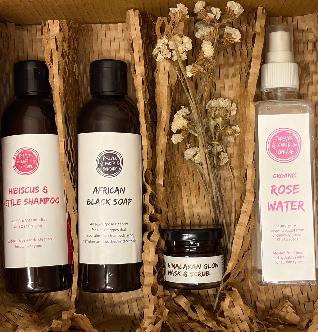 A curated gift set of four haircare products from Fresh Hair & Skin Love arranged in a woven wicker basket. The set includes a bottle of Hibiscus & Basil Shampoo, a bottle of African Black Soap, a jar of Himalayan Glow Mask & Scrub, and a spray bottle of Organic Rose Water, accented with dried white flowers.