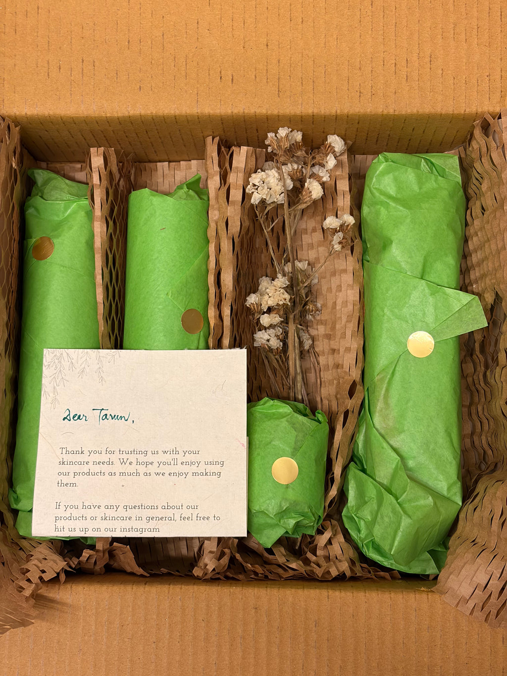 A gift box containing skincare items wrapped in bright green tissue paper with gold stickers, shredded brown paper filler, dried white flowers, and a handwritten note addressed to 'Tara'.